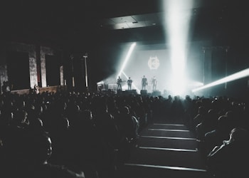 A concert venue filled with a large audience seated in rows, focused on a brightly lit stage where a band is performing with intense spotlights streaming across the scene. The atmosphere is dark and moody, with most of the light concentrated on the performers.
