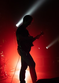 Moody silhouette of the guitarist playing against a backdrop of deep red stage lights.