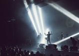 A band performing on stage with dramatic lighting and smoke effects. Bright spotlights create beams across the stage, silhouetting the musicians. A crowd is visible in the foreground, enjoying the live music.