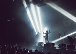 A band performing on stage with dramatic lighting and smoke effects. Bright spotlights create beams across the stage, silhouetting the musicians. A crowd is visible in the foreground, enjoying the live music.