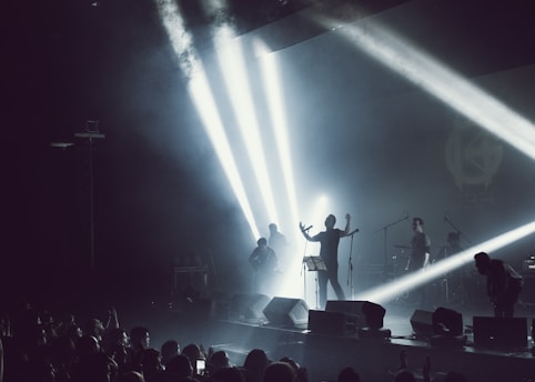 A band performing on stage with dramatic lighting and smoke effects. Bright spotlights create beams across the stage, silhouetting the musicians. A crowd is visible in the foreground, enjoying the live music.