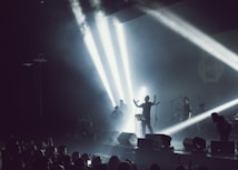 A band performing on stage with dramatic lighting and smoke effects. Bright spotlights create beams across the stage, silhouetting the musicians. A crowd is visible in the foreground, enjoying the live music.