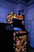An arcade game machine covered in various colorful stickers, positioned in a tiled room. The screen is glowing blue, and the machine is equipped with a red joystick and multiple buttons.
