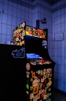 An arcade game machine covered in various colorful stickers, positioned in a tiled room. The screen is glowing blue, and the machine is equipped with a red joystick and multiple buttons.