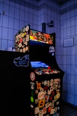 An arcade game machine covered in various colorful stickers, positioned in a tiled room. The screen is glowing blue, and the machine is equipped with a red joystick and multiple buttons.