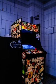 An arcade game machine covered in various colorful stickers, positioned in a tiled room. The screen is glowing blue, and the machine is equipped with a red joystick and multiple buttons.