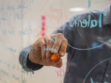 A person is writing on a clear glass or transparent board with colorful markers, including orange and blue. The writing is a mix of text and diagrams, with words like 'key', 'leadership', and 'talent'. The person's hand and a portion of their sleeve are visible, suggesting a casual setting, possibly in an office or educational environment. The focus is on the writing and the action of using the marker.