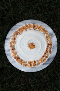 A white frosted cake is decorated with a circular arrangement of toasted almond slices on top. The cake is placed on a round marble platter, and the background consists of green grass.
