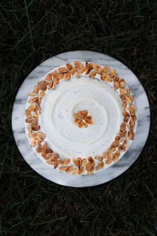 A white frosted cake is decorated with a circular arrangement of toasted almond slices on top. The cake is placed on a round marble platter, and the background consists of green grass.