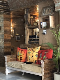A rustic living space featuring a wooden bench with colorful, decorative cushions showcasing tribal art. The walls and ceiling are made of rustic wood and clay, enhancing the earthy ambiance. The area is decorated with wooden shelves holding books and antique items. A plant adds a touch of greenery to the setting, and framed pictures adorn the walls.