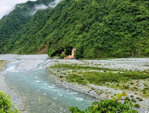A serene landscape featuring lush green mountains with mist gently hugging the peaks. A small river flows through the valley, bordered by rocky banks and verdant vegetation. Tucked amidst the greenery is a picturesque temple with bright, earthy tones, providing a striking contrast to its natural surroundings.