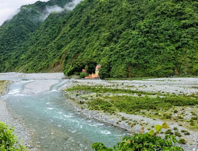 A serene landscape featuring lush green mountains with mist gently hugging the peaks. A small river flows through the valley, bordered by rocky banks and verdant vegetation. Tucked amidst the greenery is a picturesque temple with bright, earthy tones, providing a striking contrast to its natural surroundings.