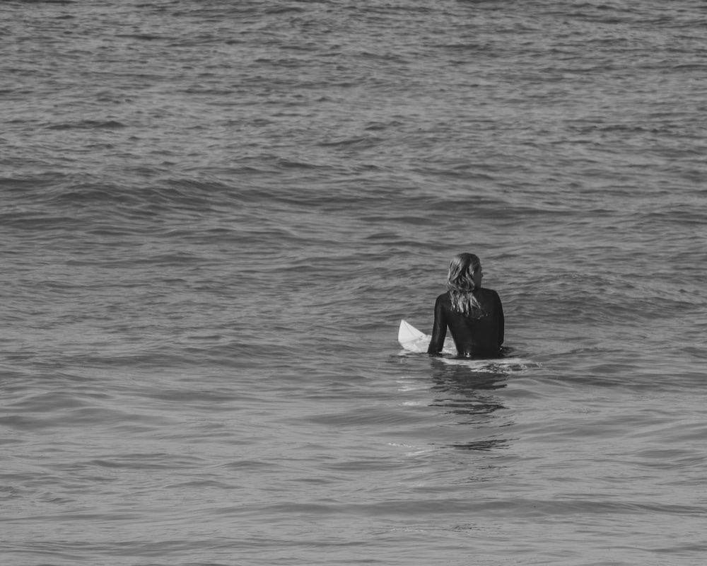 grayscale photo of woman in black wetsuit sitting on surfboard on water