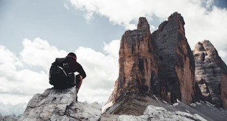 Hiker resting on a boulder, gazing at the expansive mountain vista under cloudy skies.