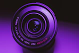 Close-up of a sleek camera lens reflecting vibrant purple light.
