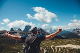 A person with a backpack stands on a mountain path, arms outstretched, facing a stunning mountainous landscape. The sky is partly cloudy, and the scene conveys a sense of freedom and adventure.