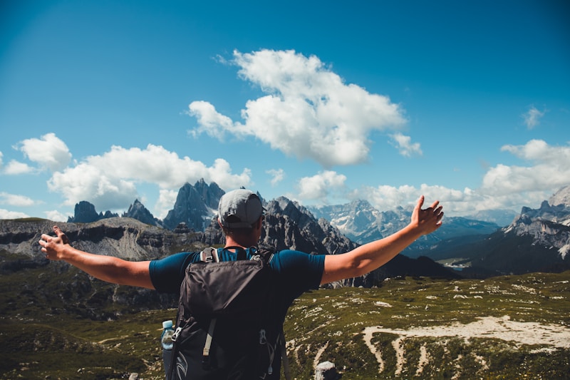 A person with a backpack stands on a mountain path, arms outstretched, facing a stunning mountainous landscape. The sky is partly cloudy, and the scene conveys a sense of freedom and adventure.