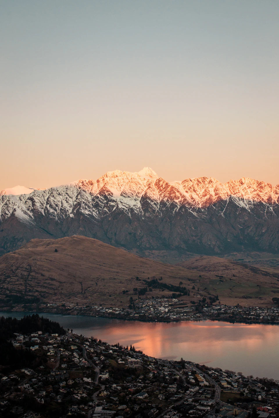 The image depicts a breathtaking view of a mountain range at sunset, with the sun casting its warm glow on the snow-capped peaks and illuminating the surrounding landscape. The mountains are bathed in hues of orange and yellow, creating a stunning contrast against the deep blue sky. Below, a town nestles comfortably amidst the natural beauty, with houses and buildings scattered across the scene. The image is taken from an elevated perspective, providing a comprehensive view of the majestic mountain range and its picturesque setting.