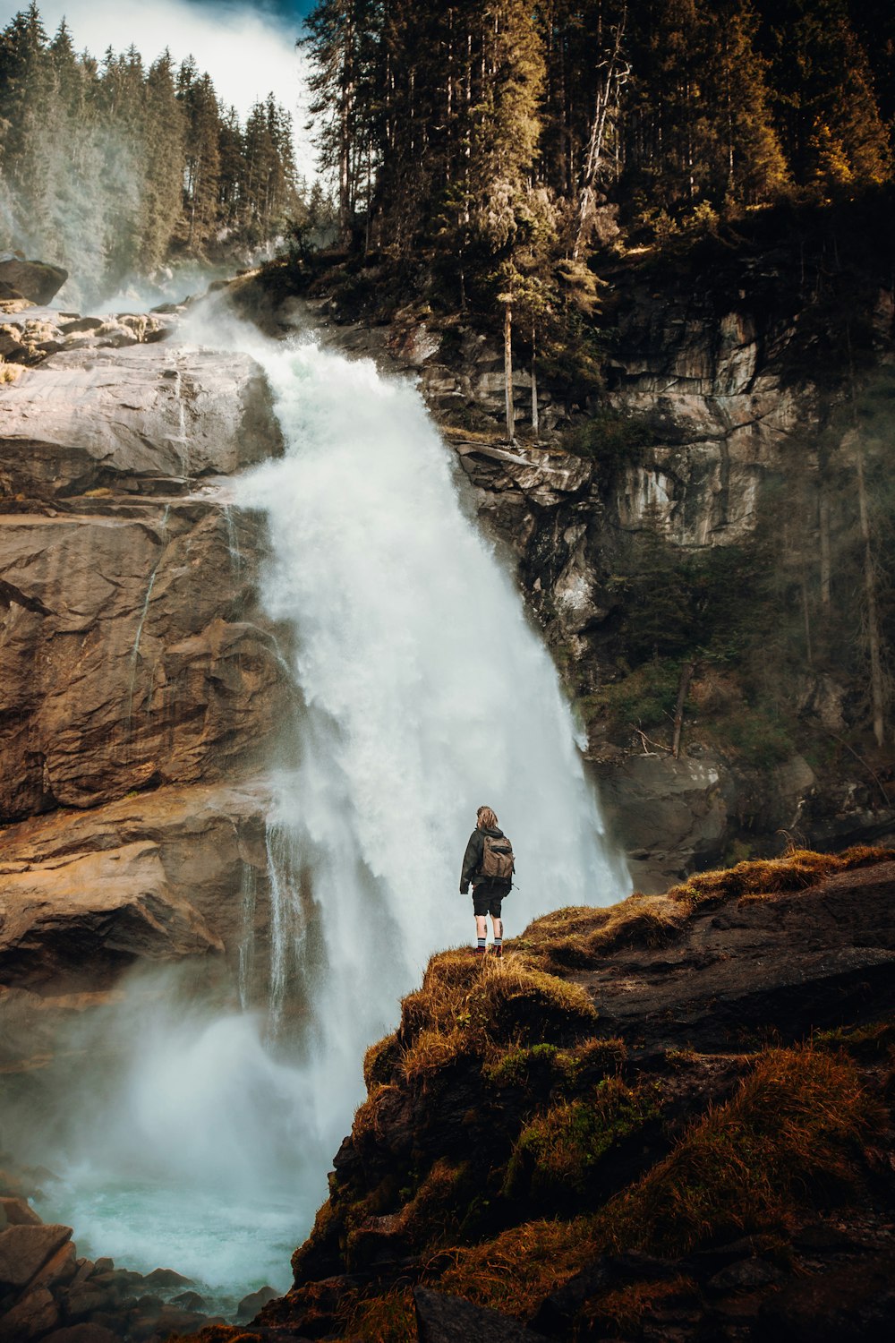 昼間、滝の近くの茶色の岩の上に立つ黒いジャケットの男