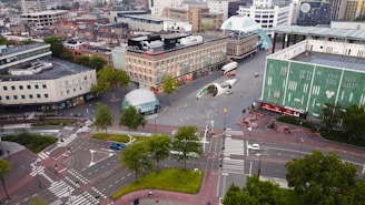 Aerial view of an urban area featuring a large, open square surrounded by modern and mid-century buildings. There are roads with several cars and bicycles, crosswalks, greenery, and public walkways. Notable objects include a spherical glass structure, a futuristic archway, and various trees. Pedestrians and vehicles are visible, adding to the city's busy atmosphere.