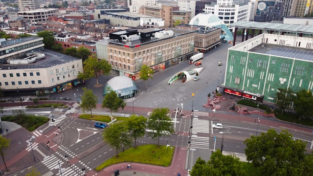 Aerial view of an urban area featuring a large, open square surrounded by modern and mid-century buildings. There are roads with several cars and bicycles, crosswalks, greenery, and public walkways. Notable objects include a spherical glass structure, a futuristic archway, and various trees. Pedestrians and vehicles are visible, adding to the city's busy atmosphere.