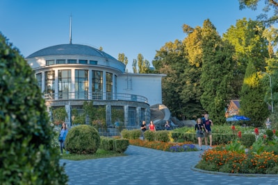 A circular building with large windows is situated in a lush garden setting with neatly trimmed bushes and colorful flowers. People are casually walking around the pathway, enjoying the outdoor space. Tall trees frame the scene, providing a serene and natural atmosphere.