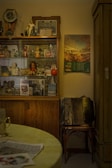 A vintage room corner featuring a glass cabinet filled with various small figurines, photographs, and decorative items. A 1985 calendar hangs on the wall beside a wooden cabinet. An old accordion sits on a chair next to the cabinet. The setup exudes a nostalgic atmosphere with a mix of old and personal memorabilia. A small table with a light-colored tablecloth is partially visible in the foreground.
