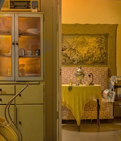 A vintage kitchen with an old radio placed on top of a weathered cabinet. Plates and a clock are visible through the glass doors. On the right, a cozy room displays a tapestry with a picturesque scene, and a samovar sits on a table draped with a yellow cloth, surrounded by ornate wallpaper and textiles.