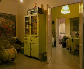 A cozy vintage living space featuring an old cabinet with glass doors filled with various objects. A lampshade emits warm light, and a tapestry depicting nature scenes decorates the wall. A worn-looking armchair and an antique pram are also present. The open door leads to a second, similarly decorated room with a table covered with a tablecloth, a samovar, and chairs.