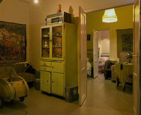 A cozy vintage living space featuring an old cabinet with glass doors filled with various objects. A lampshade emits warm light, and a tapestry depicting nature scenes decorates the wall. A worn-looking armchair and an antique pram are also present. The open door leads to a second, similarly decorated room with a table covered with a tablecloth, a samovar, and chairs.