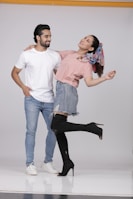 A man and a woman are posing playfully in casual outfits. The man is wearing a white t-shirt and jeans, while the woman is dressed in a pink top, denim skirt, and black high-heeled boots. She has a colorful scarf tied in her hair and is playfully leaning back with one leg raised.