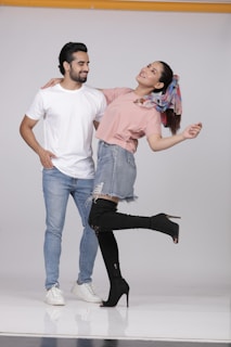 A man and a woman are posing playfully in casual outfits. The man is wearing a white t-shirt and jeans, while the woman is dressed in a pink top, denim skirt, and black high-heeled boots. She has a colorful scarf tied in her hair and is playfully leaning back with one leg raised.