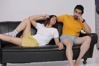 A man and a woman are relaxing on a black couch. The woman, wearing a white t-shirt and yellow shorts, is lying down with her head tilted back and a playful expression. The man, wearing a yellow t-shirt and light blue shorts, is sitting with a thoughtful pose and a slight smile. The background is plain and minimalistic.