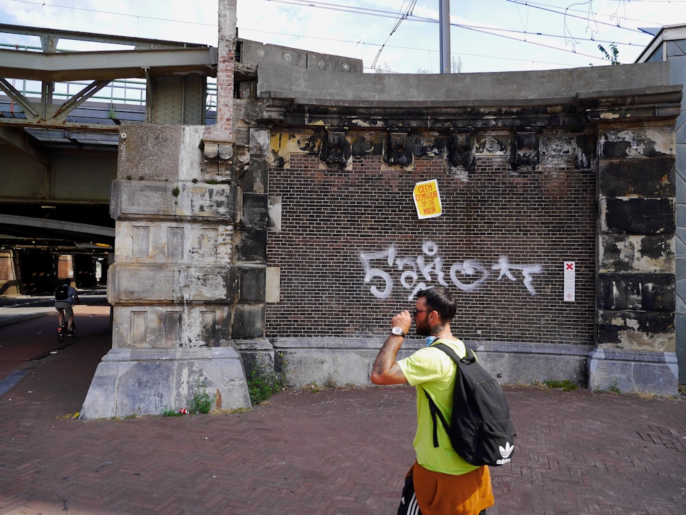 黄色いシャツを着て灰色のリュックサックを背負った男が、昼間、灰色のコンクリートの壁のそばに立っている
