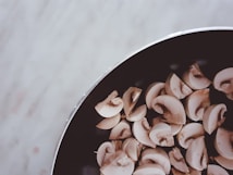 A collection of sliced mushrooms is placed inside a dark pan, positioned on the right side of the image. The backdrop features a light, textured surface, providing a contrast to the darker tones of the pan.