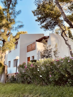 A two-story white building with wooden accents is partially surrounded by lush greenery and colorful flowering shrubs. Tall trees frame the setting, casting gentle shadows against the structure. The sky is clear and blue, enhancing the tranquil ambiance.