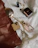 A collection of luxury items is laid out on a soft white fabric. The items include a woven brown leather bag, a smartphone with a gold finish, a bottle of perfume with a honeycomb design cap, a compact makeup item, and a black lipstick. There is also a gold chain with a pendant. The setup gives a sense of elegance and sophistication.