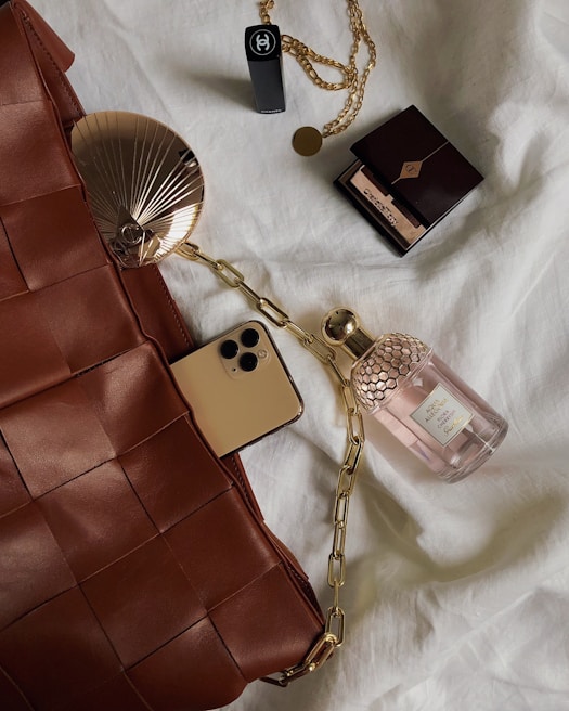 A collection of luxury items is laid out on a soft white fabric. The items include a woven brown leather bag, a smartphone with a gold finish, a bottle of perfume with a honeycomb design cap, a compact makeup item, and a black lipstick. There is also a gold chain with a pendant. The setup gives a sense of elegance and sophistication.