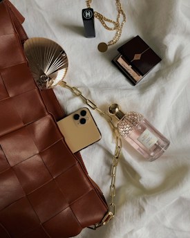 A collection of luxury items is laid out on a soft white fabric. The items include a woven brown leather bag, a smartphone with a gold finish, a bottle of perfume with a honeycomb design cap, a compact makeup item, and a black lipstick. There is also a gold chain with a pendant. The setup gives a sense of elegance and sophistication.