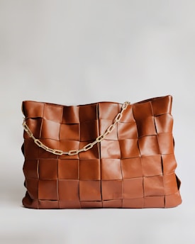 A large handbag made of woven leather squares, featuring a brown hue and a gold chain strap. The bag's texture and design give it a sophisticated and elegant appearance.