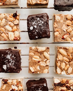 A close-up view of several small square dessert bars arranged on a rack. The bars appear to have a layer of chocolate topped with coarse sea salt, while others are adorned with a generous amount of nuts. The texture of the desserts indicates a mix of chocolate and nutty toppings.