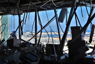 A chaotic scene with broken and tangled metal structures and wires in the foreground. The background shows a port area with debris scattered around, suggesting damage or destruction. There is a view of the sea and partially damaged buildings visible through the wreckage.