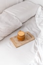 A cozy setting featuring a neatly made bed with white linens. On the bed lies a wooden cutting board holding a mustard-yellow ceramic mug with a handle. The image conveys a sense of tranquility and comfort.