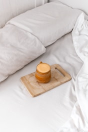 A cozy setting featuring a neatly made bed with white linens. On the bed lies a wooden cutting board holding a mustard-yellow ceramic mug with a handle. The image conveys a sense of tranquility and comfort.