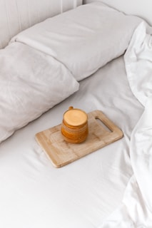 A cozy setting featuring a neatly made bed with white linens. On the bed lies a wooden cutting board holding a mustard-yellow ceramic mug with a handle. The image conveys a sense of tranquility and comfort.