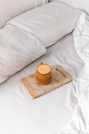 A cozy setting featuring a neatly made bed with white linens. On the bed lies a wooden cutting board holding a mustard-yellow ceramic mug with a handle. The image conveys a sense of tranquility and comfort.