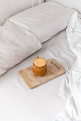 A cozy setting featuring a neatly made bed with white linens. On the bed lies a wooden cutting board holding a mustard-yellow ceramic mug with a handle. The image conveys a sense of tranquility and comfort.