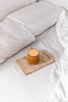 A cozy setting featuring a neatly made bed with white linens. On the bed lies a wooden cutting board holding a mustard-yellow ceramic mug with a handle. The image conveys a sense of tranquility and comfort.