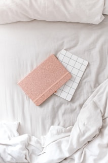 A cozy, minimalist setting features two notebooks placed on a soft, white bedspread. One notebook has a sparkly rose gold cover, while the other has a white cover with a black grid pattern. The sheets are slightly rumpled, creating a relaxed atmosphere.