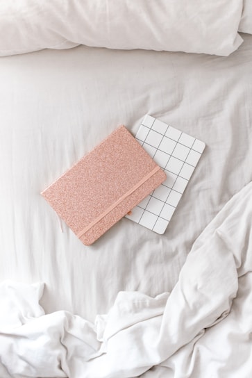 A cozy, minimalist setting features two notebooks placed on a soft, white bedspread. One notebook has a sparkly rose gold cover, while the other has a white cover with a black grid pattern. The sheets are slightly rumpled, creating a relaxed atmosphere.